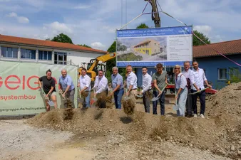 Eine Gruppe von Menschen mit Schaufeln posiert bei einer Grundsteinlegung, im Hintergrund Baugeräte und Schilder, an einem sonnigen Tag.