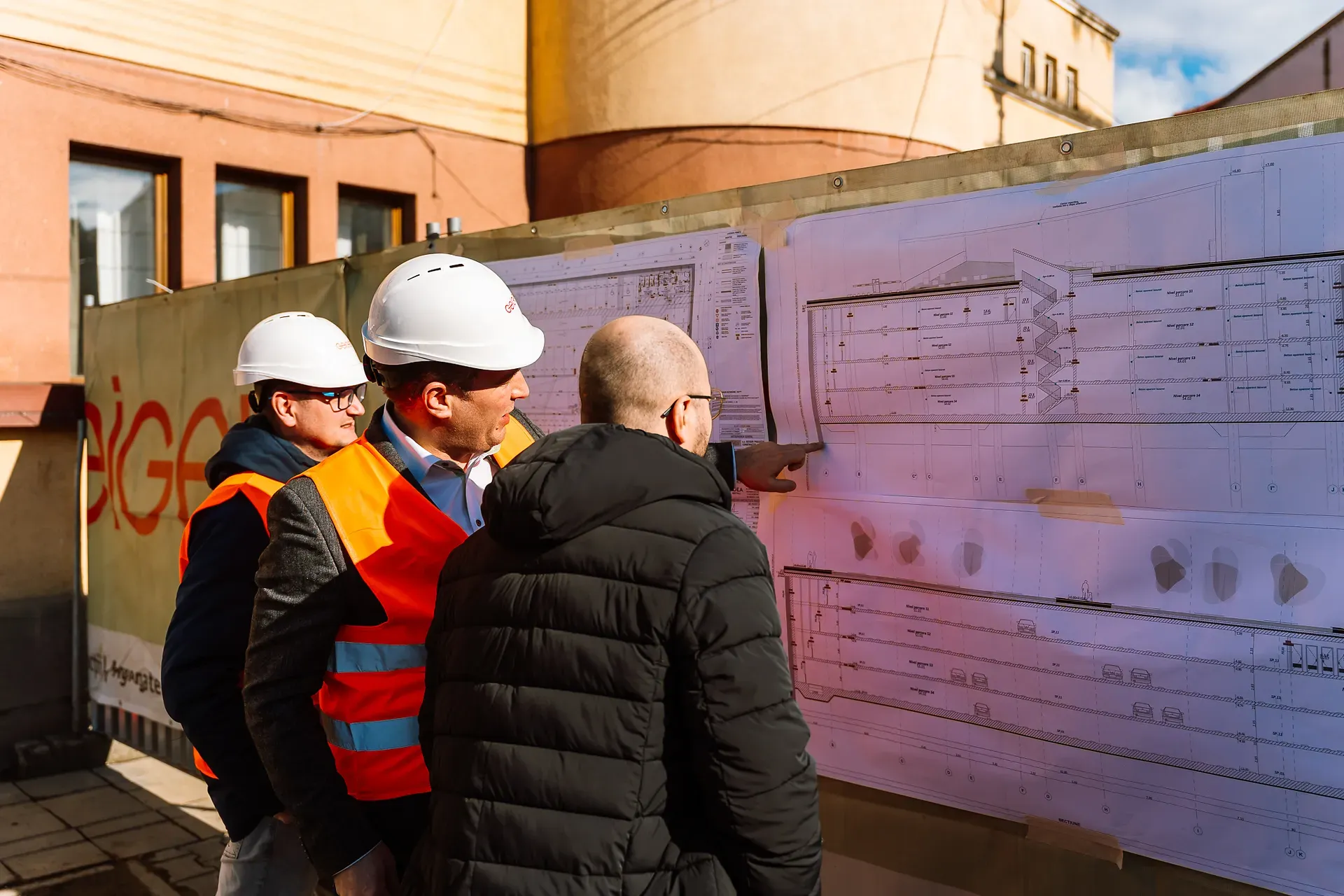 Trei muncitori în construcții cu căști și veste de protecție examinează planuri pe un perete la un șantier însorit.