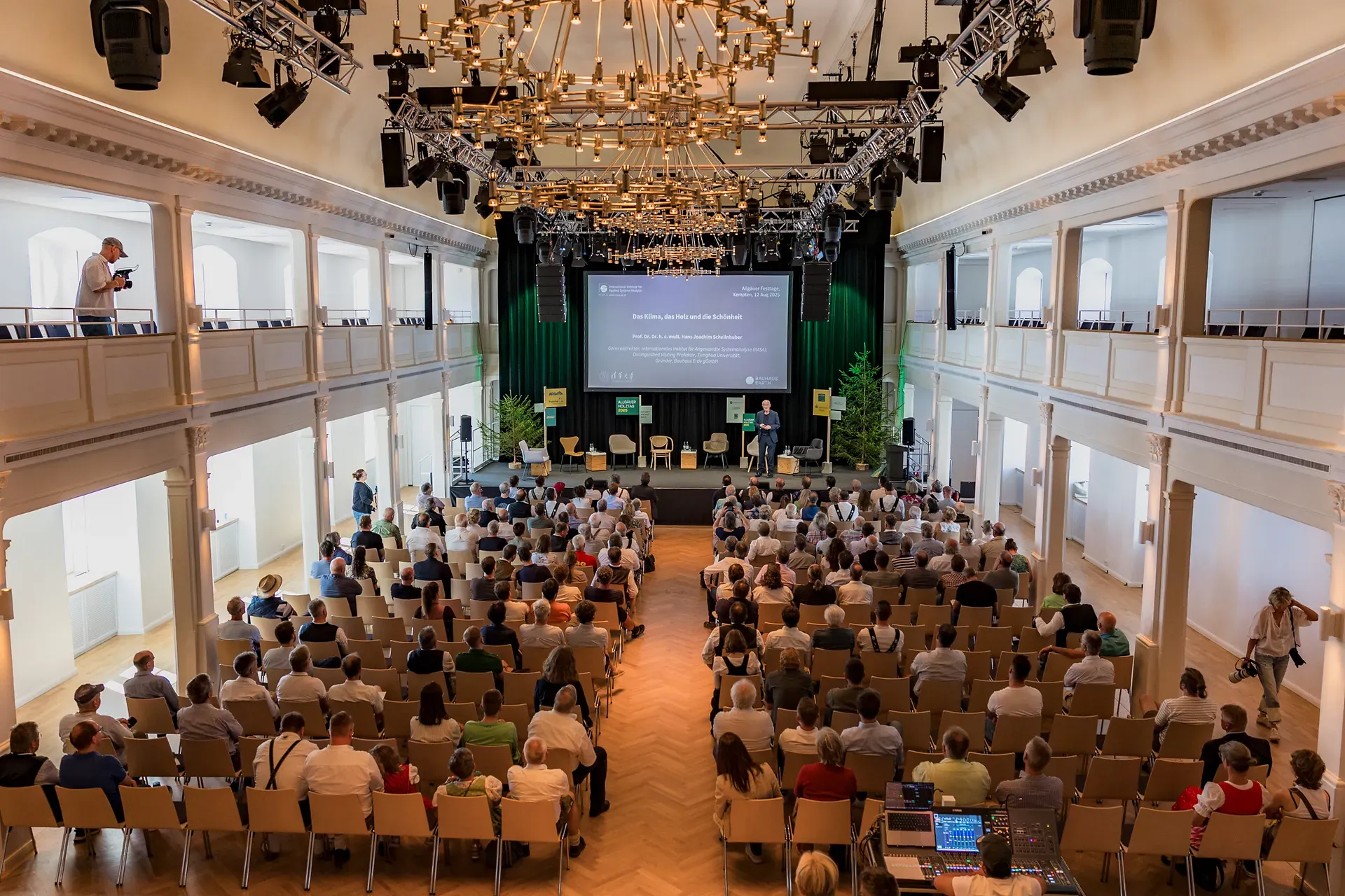 Ein großer Konferenzraum beim Holztag 2025 in Kempten mit einem Publikum vor einer Bühne mit Präsentationsbildschirm und Lautsprechern. Der Raum verfügt über elegante Kronleuchter und Balkone.