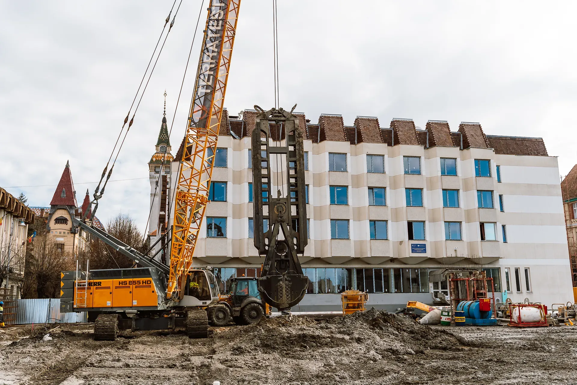 Șantier mare de construcții cu un macarale și utilaje în fața unei clădiri moderne într-o zi înnorată. Arhitectură vintage este vizibilă în fundal.