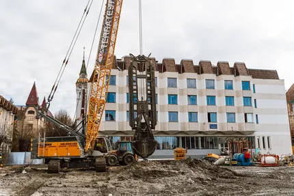 Șantier mare de construcții cu un macarale și utilaje în fața unei clădiri moderne într-o zi înnorată. Arhitectură vintage este vizibilă în fundal.