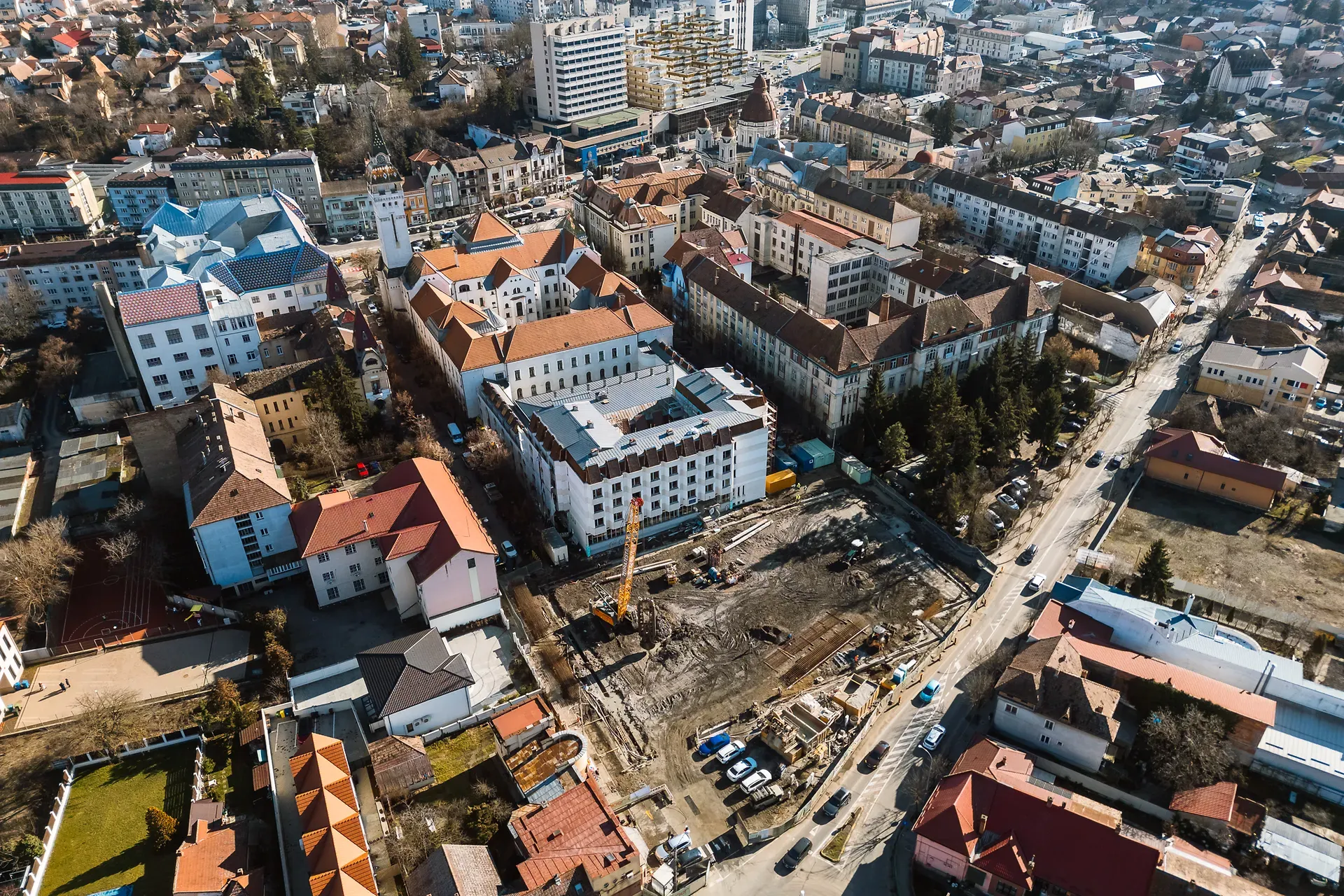 Vedere aeriană a unui oraș cu clădiri cu acoperișuri portocalii, șantier de construcții, vehicule și peisaj urban înconjurător sub un cer senin.