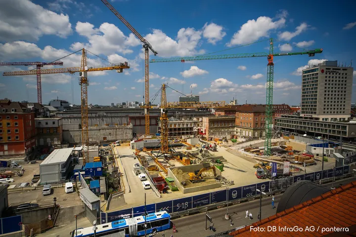Baustelle mit mehreren Kränen, umgeben von Gebäuden unter einem blauen Himmel mit Wolken. Stadtentwicklung im Gange.