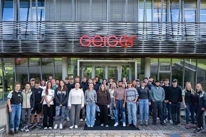 Eine Gruppe von neuen Auszubildenden steht vor dem Standort Oberstdorf der Geiger Gruppe,  mit dem Schriftzug „Geiger“ über dem Eingang und posiert für ein Gruppenfoto.