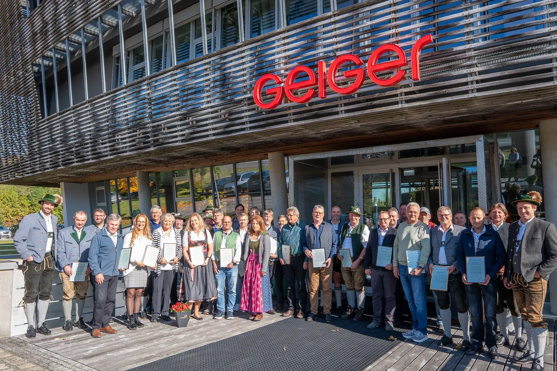 Jubilare der Geiger Gruppe in traditioneller Tracht vor dem Firmengebäude mit Geiger-Schriftzug, halten Urkunden in den Händen und lächeln in die Kamera.
