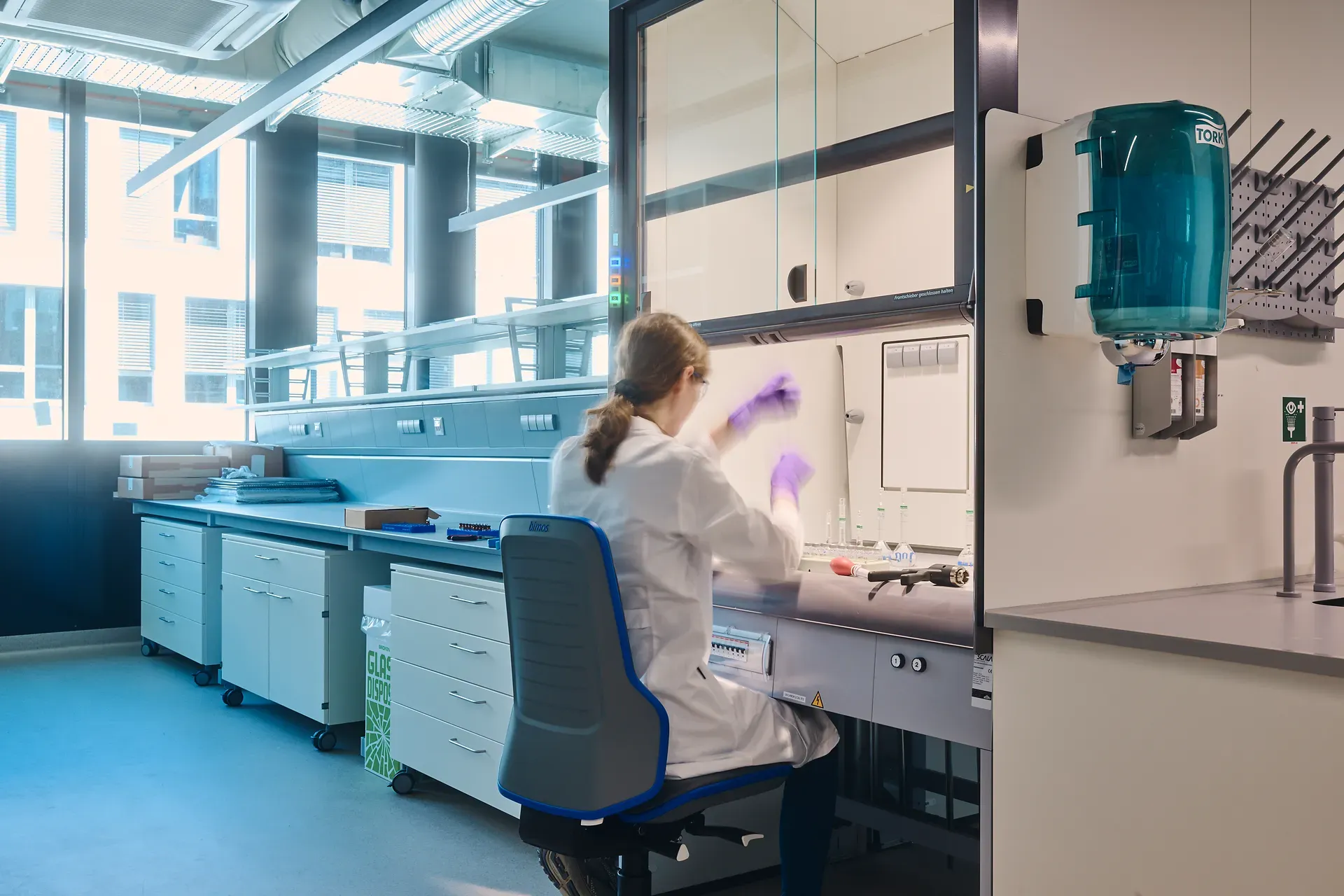Wissenschaftler im Laborkittel und mit lila Handschuhen arbeitet an einem Labortisch, umgeben von Geräten und Lagerschränken.