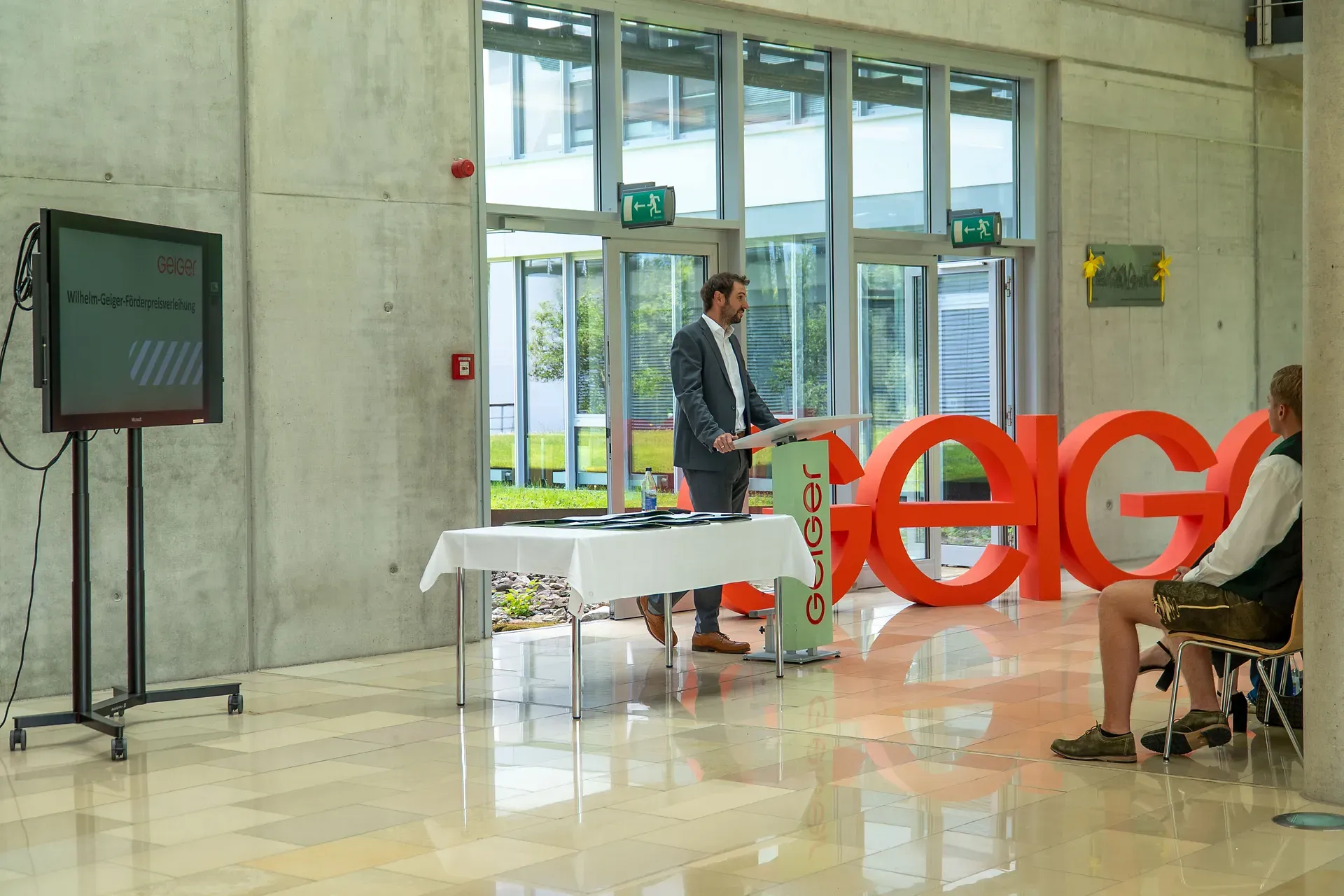 Ein Mann im Anzug spricht an einem Podium in einem modernen Gebäude. Auf einem Bildschirm ist „Geiger“ zu sehen und in der Nähe stehen große rote Buchstaben, die „Geiger“ buchstabieren.