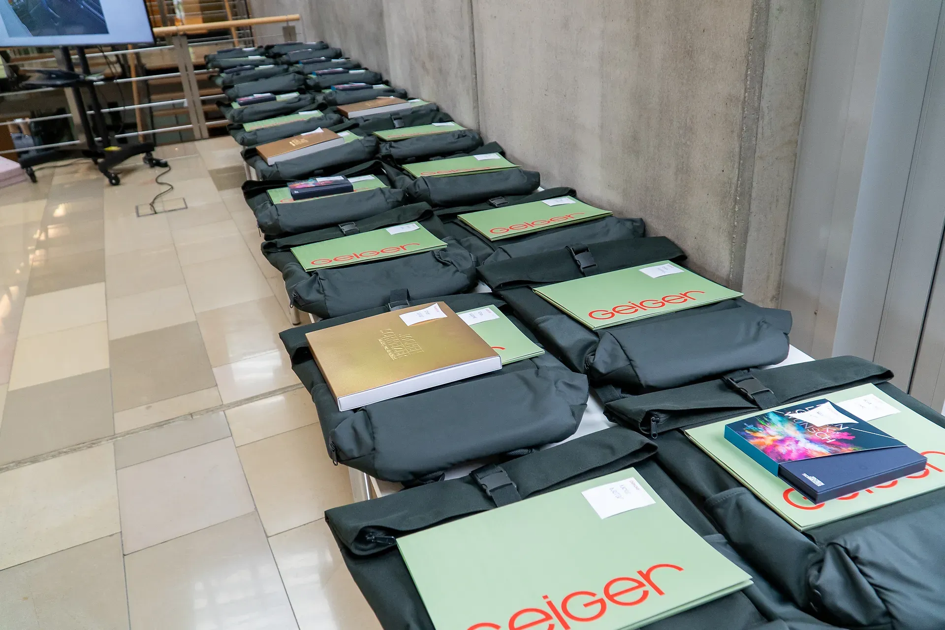 Reihen schwarzer Konferenztaschen mit Ordnern, Notizbüchern und Tablets auf einem gefliesten Boden vor einer Betonwand.