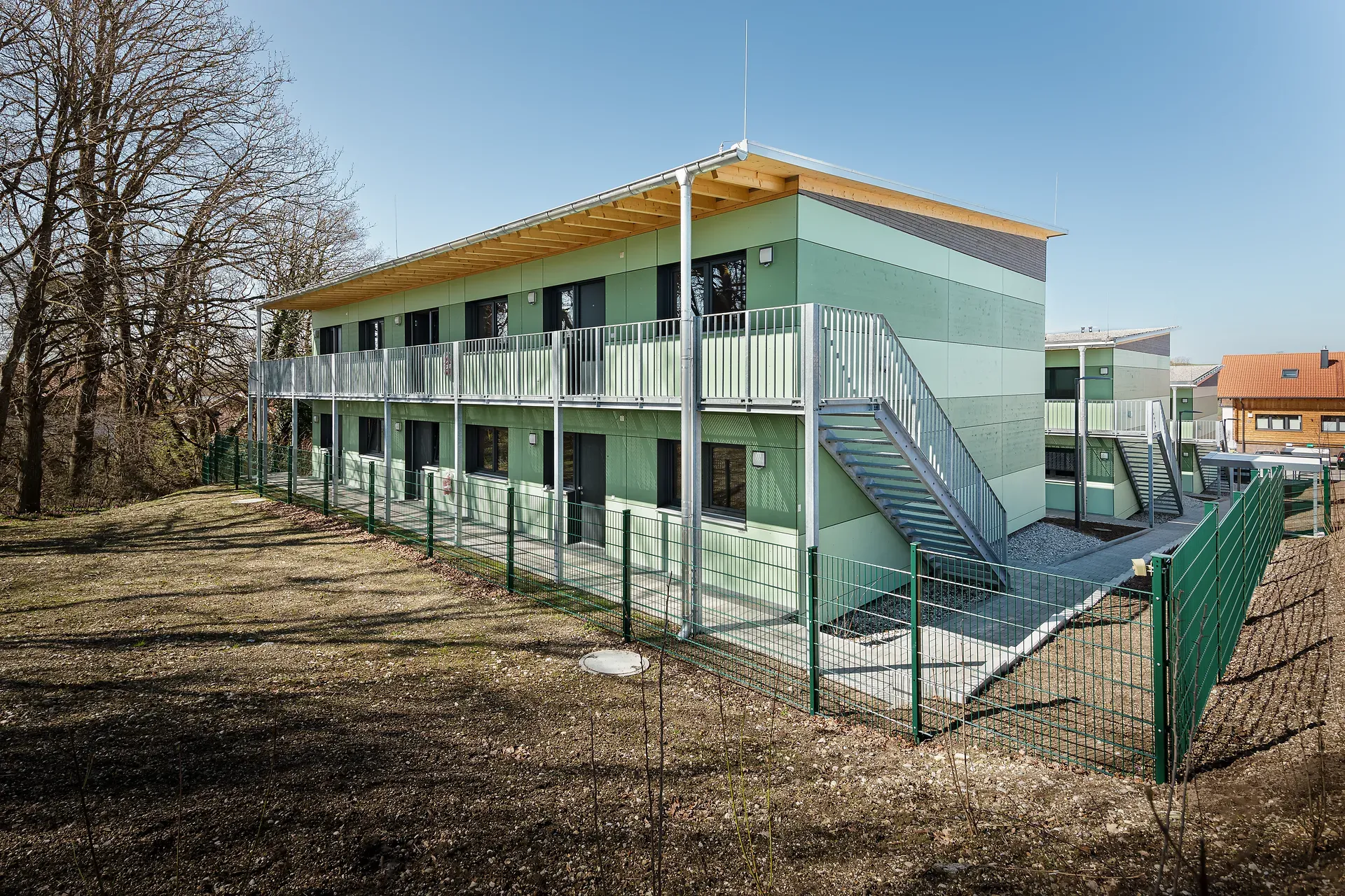 Modernes zweistöckiges grünes Gebäude mit Balkonen, Außentreppen und eingezäuntem Hof, umgeben von kahlen Bäumen unter klarem blauem Himmel.