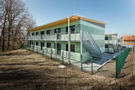 Modernes zweistöckiges grünes Gebäude mit Balkonen, Außentreppen und eingezäuntem Hof, umgeben von kahlen Bäumen unter klarem blauem Himmel.