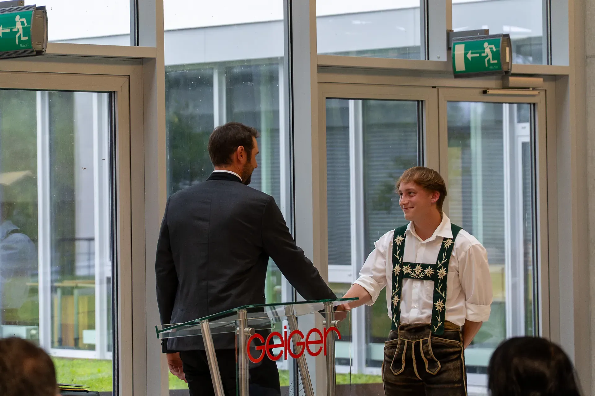 Ein Auszubildender und Pius Geiger 3 schütteln sich an einem Podium im Innenbereich die Hände. Einer trägt einen Anzug, der andere traditionelle Kleidung. Im Hintergrund sind Glastüren und Ausgangsschilder zu sehen.