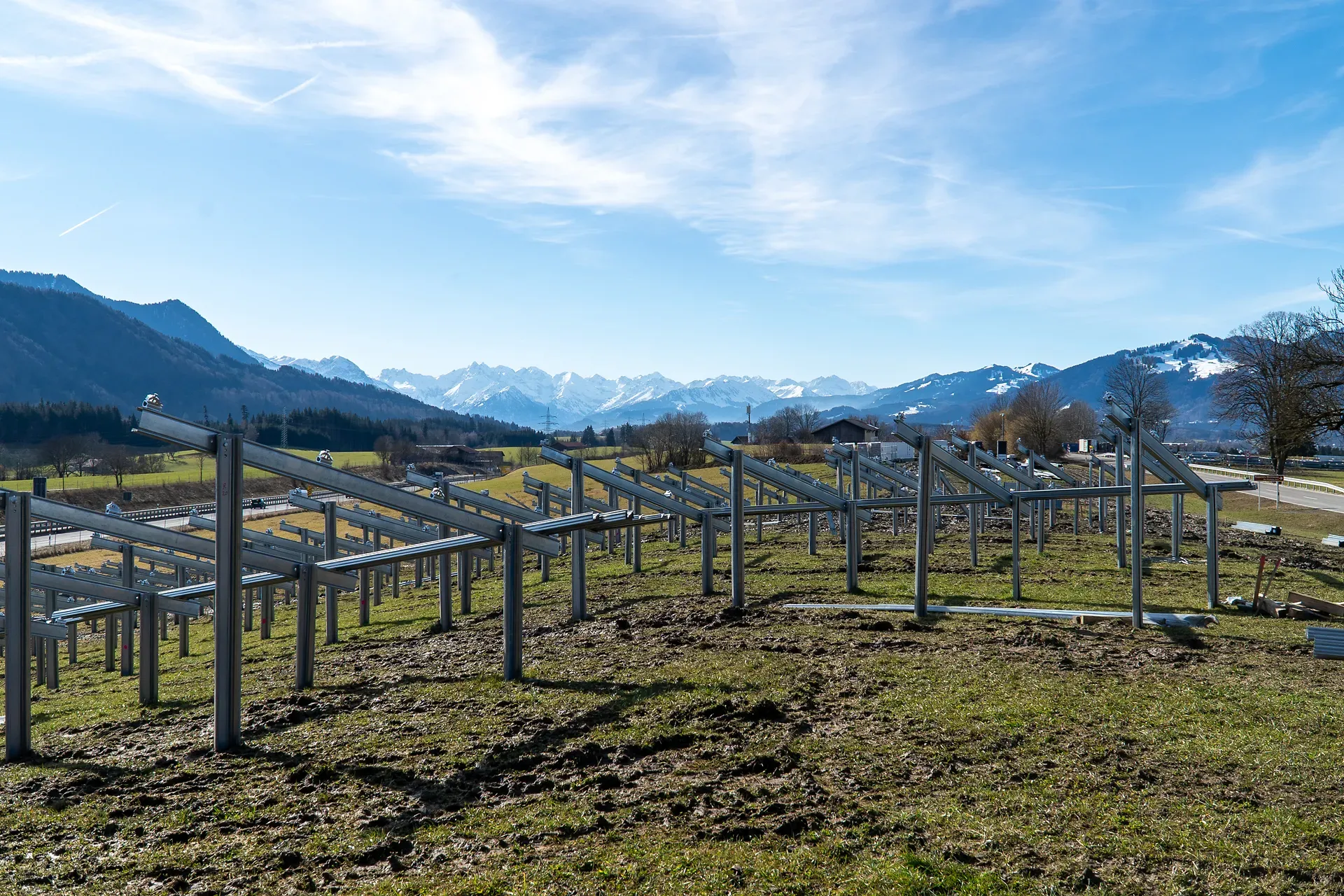 Metallrahmen für Solarmodule auf einem grasbewachsenen Hügel unter blauem Himmel, mit Bergen im Hintergrund.