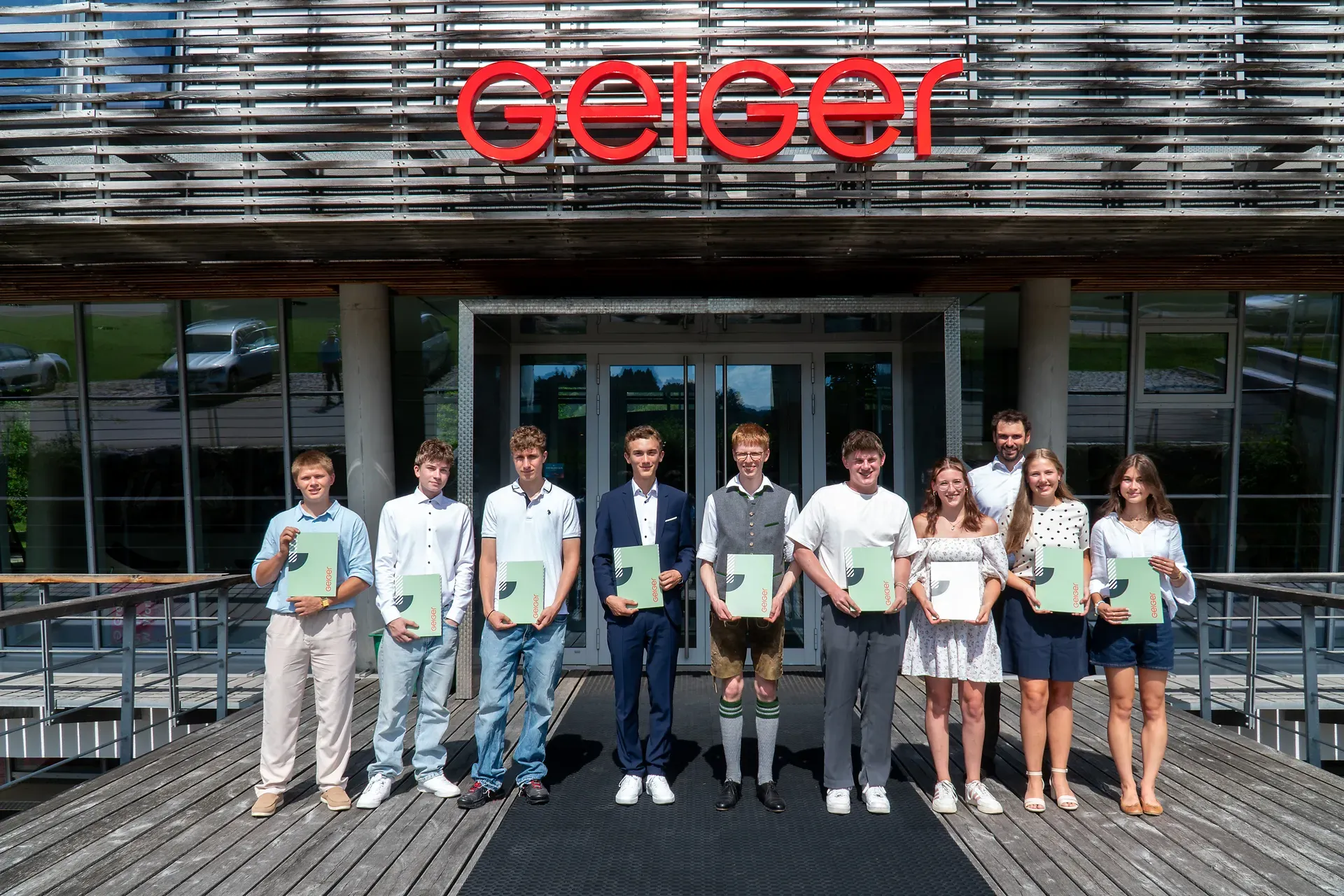 Neun Personen stehen vor einem Gebäude mit dem Schild „Geiger“ und halten Zertifikate. Sie tragen Sommerkleidung.