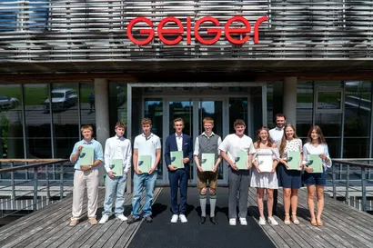 Neun Personen stehen vor einem Gebäude mit dem Schild „Geiger“ und halten Zertifikate. Sie tragen Sommerkleidung.