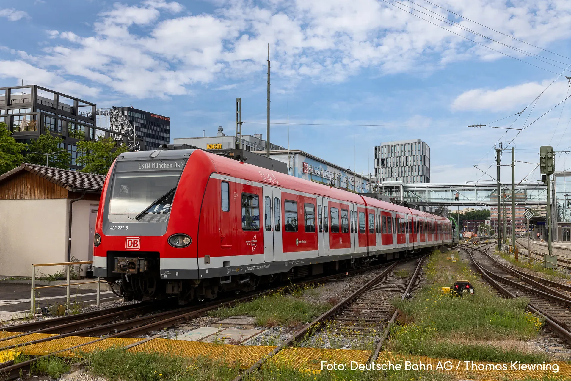 Ein roter Zug der Deutschen Bahn fährt unter einem teilweise bewölkten Himmel auf Gleisen in der Nähe moderner Gebäude.