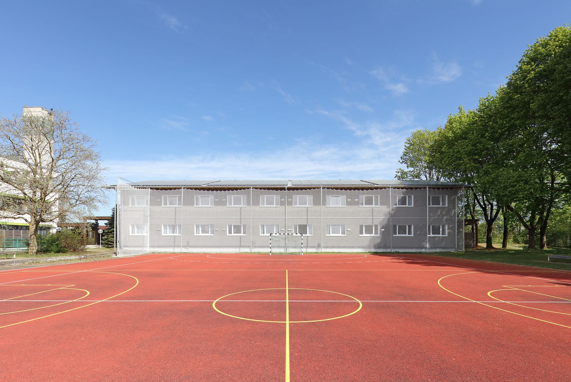 Zweistöckiger Holzmodulbau, im Vordergrund ein neuer Sportplatz mit Markierungen.