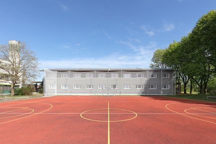 Zweistöckiger Holzmodulbau, im Vordergrund ein neuer Sportplatz mit Markierungen.
