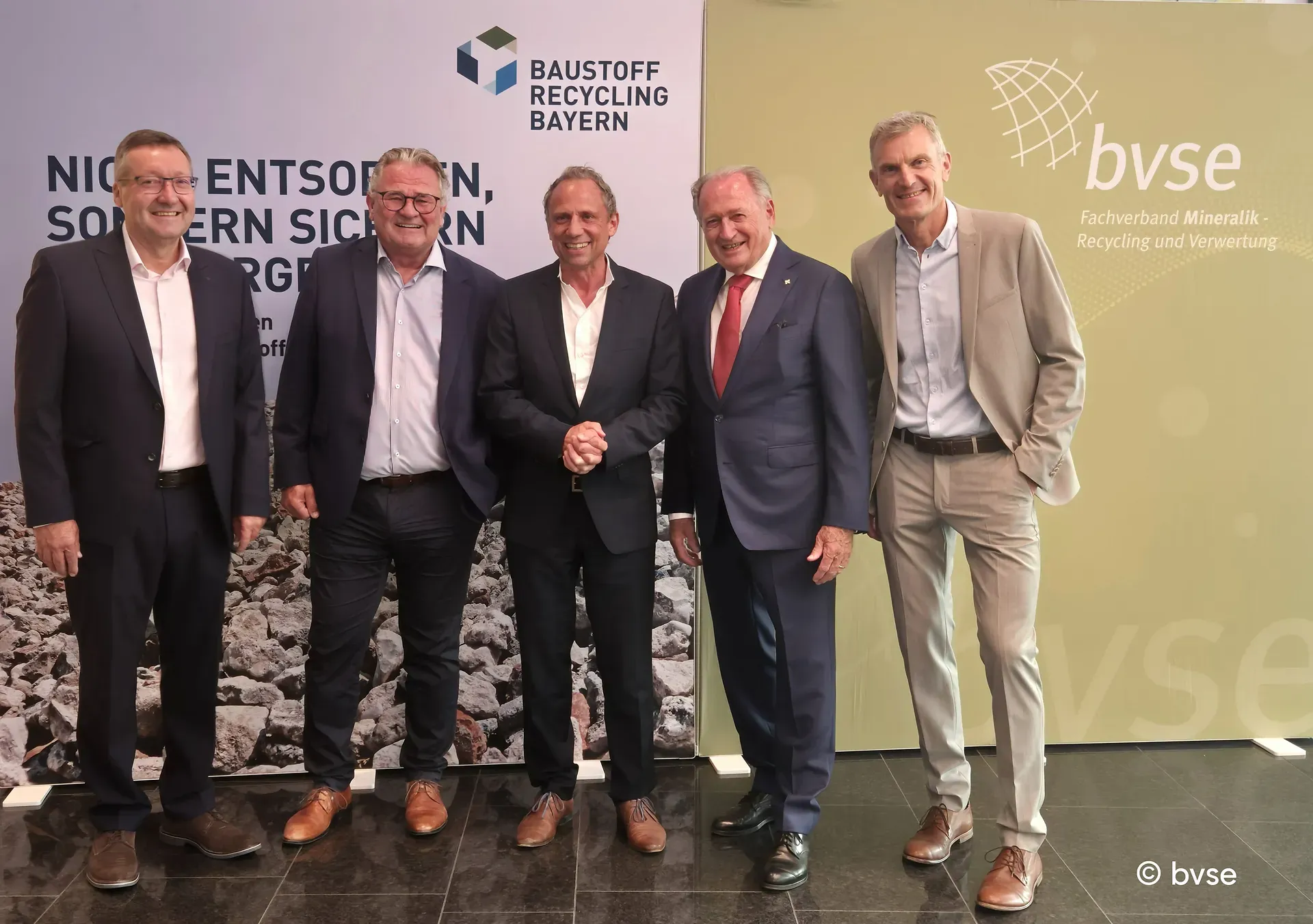 Fünf Männer in Anzügen stehen lächelnd zusammen vor Bannern von Baustoff Recycling Bayern und dem bvse.