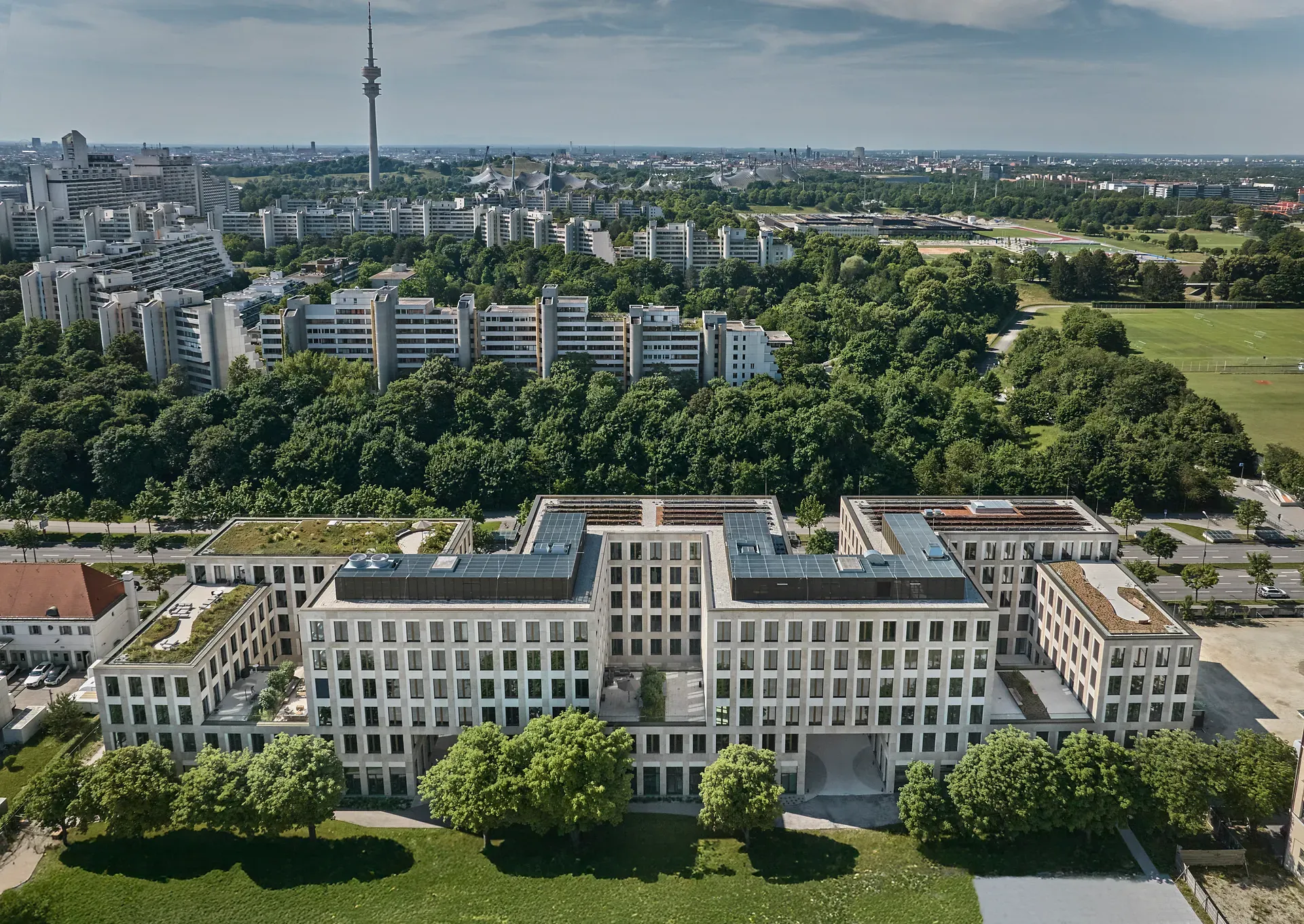 Luftaufnahme eines modernen, U-förmigen Gebäudes, umgeben von Bäumen, mit einer Stadtlandschaft und einem hohen Turm im Hintergrund unter einem klaren Himmel.