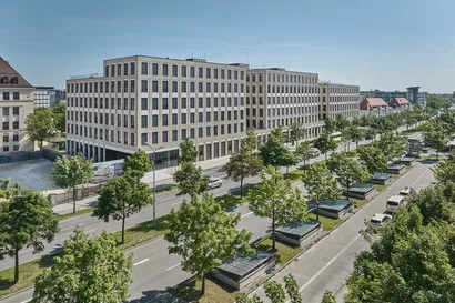 Luftaufnahme eines modernen, mehrstöckigen Bürogebäudes mit großen Fenstern, umgeben von Bäumen und einer belebten Straße mit Autos.