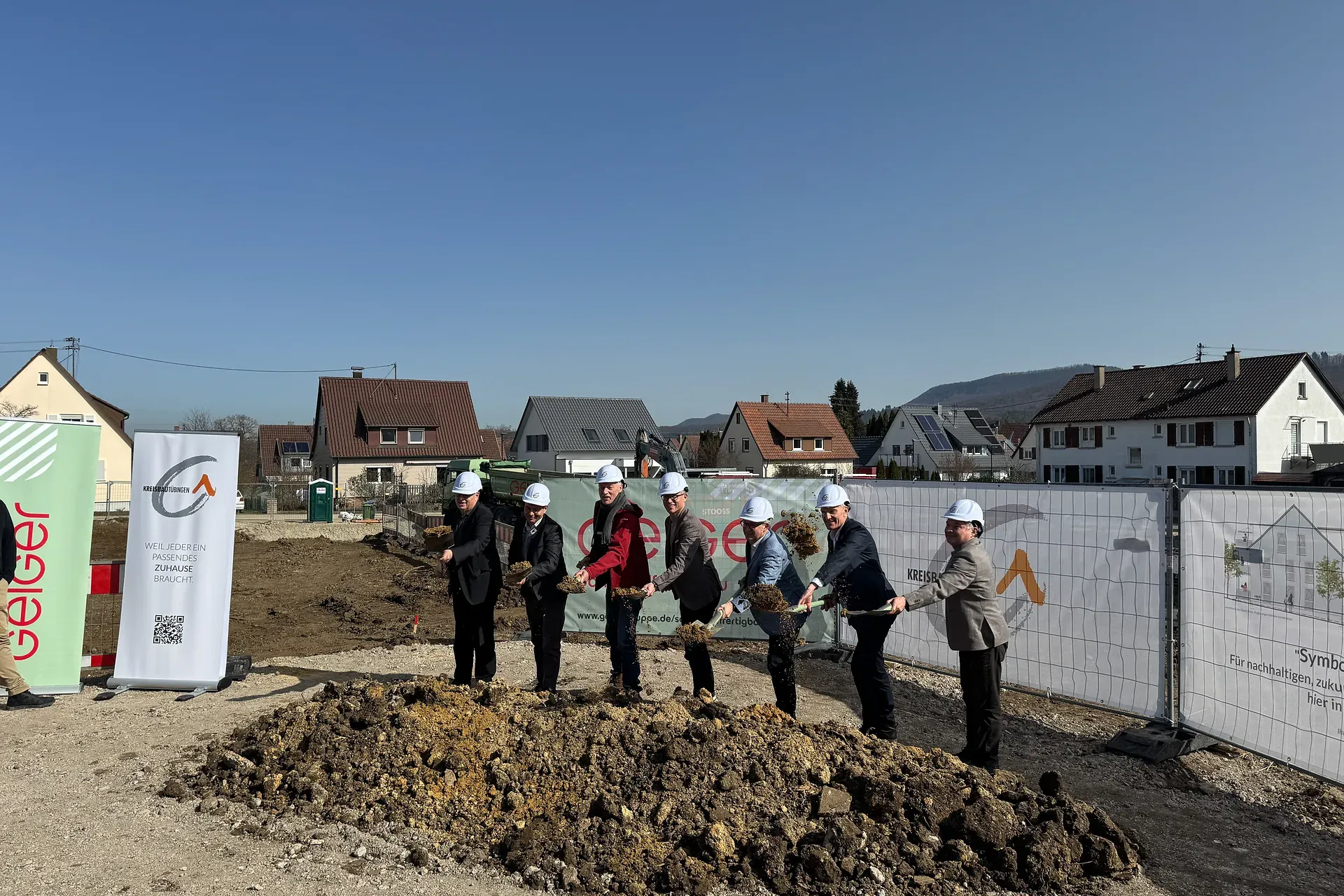Sieben Personen mit Schutzhelmen nehmen an einem Spatenstich teil und halten Schaufeln in der Nähe eines Erdbergs, mit Häusern im Hintergrund.