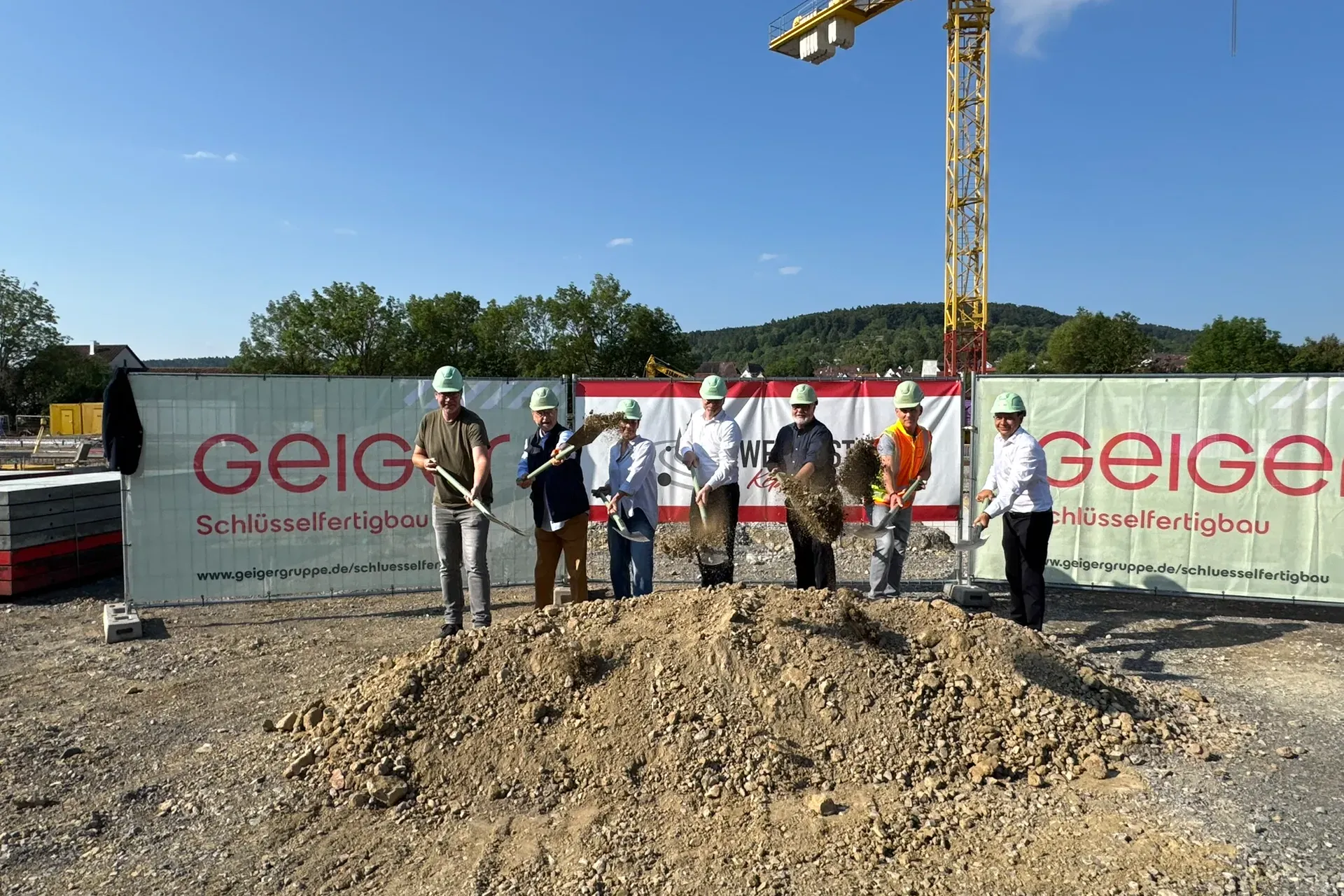 Gruppe von Personen mit Schutzhelmen und Spaten bei einem feierlichen Spatenstich auf einer Baustelle. Im Hintergrund sind ein Kran sowie Banner zu sehen.