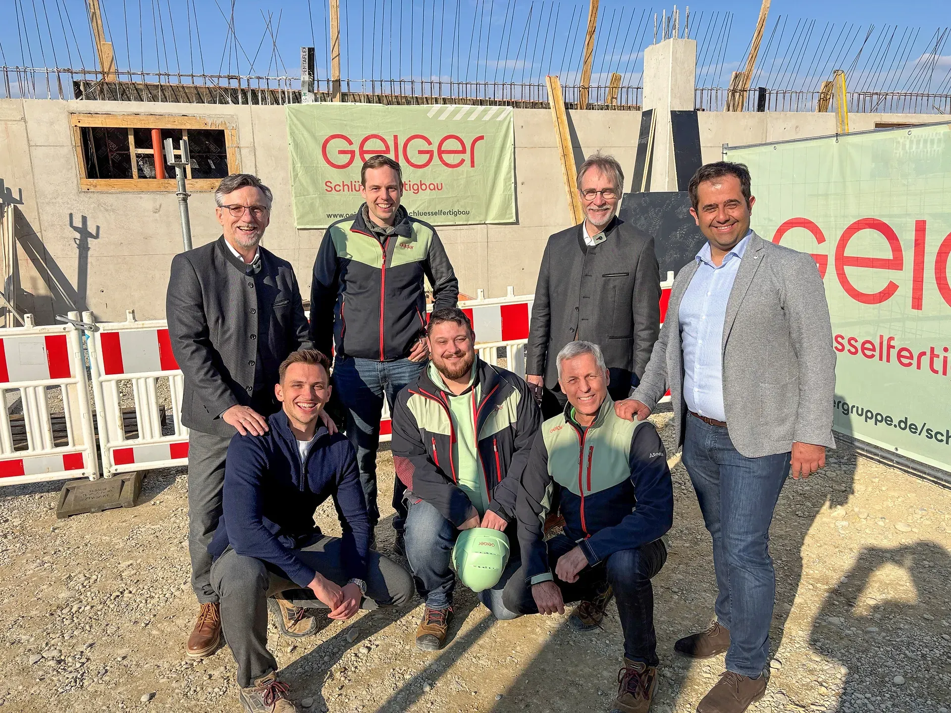 Sieben Männer posieren im Freien auf einer Baustelle mit Holzstrukturen und Geiger-Schildern unter klarem blauem Himmel.