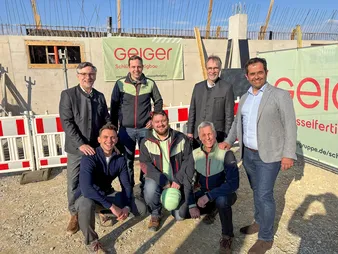Sieben Männer posieren im Freien auf einer Baustelle mit Holzstrukturen und Geiger-Schildern unter klarem blauem Himmel.