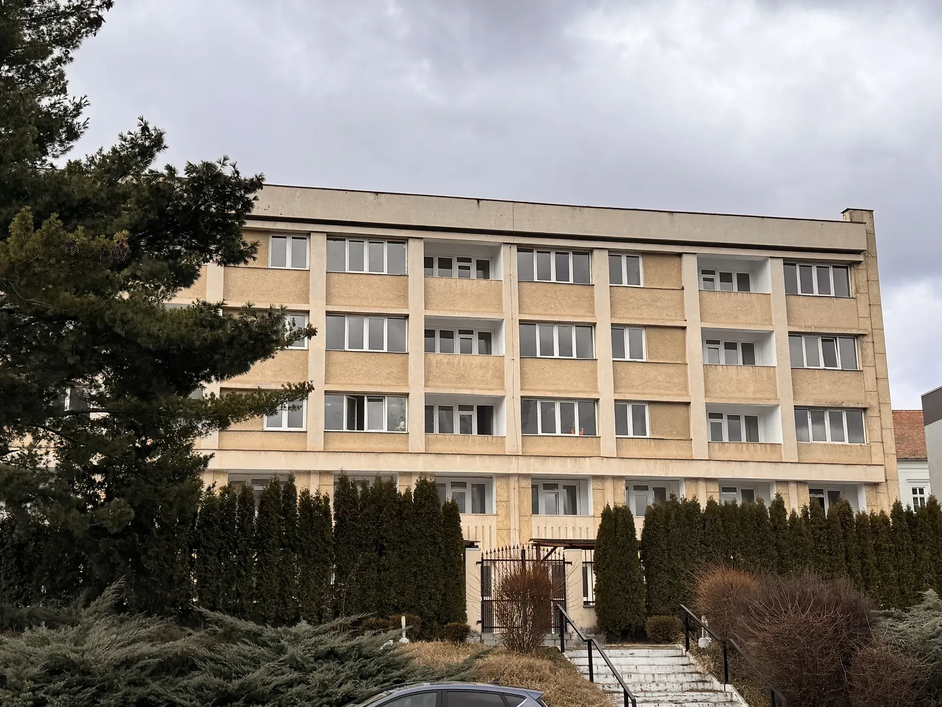 Clădire bej cu patru etaje, cu mai multe ferestre și balcoane, înconjurată de arbuști înalți și un copac, sub un cer înnorat.