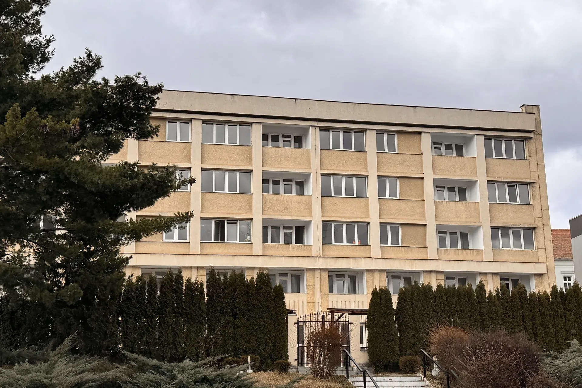 Clădire bej cu patru etaje, cu mai multe ferestre și balcoane, înconjurată de arbuști înalți și un copac, sub un cer înnorat.