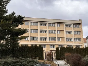 Clădire bej cu patru etaje, cu mai multe ferestre și balcoane, înconjurată de arbuști înalți și un copac, sub un cer înnorat.