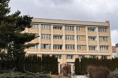 Clădire bej cu patru etaje, cu mai multe ferestre și balcoane, înconjurată de arbuști înalți și un copac, sub un cer înnorat.