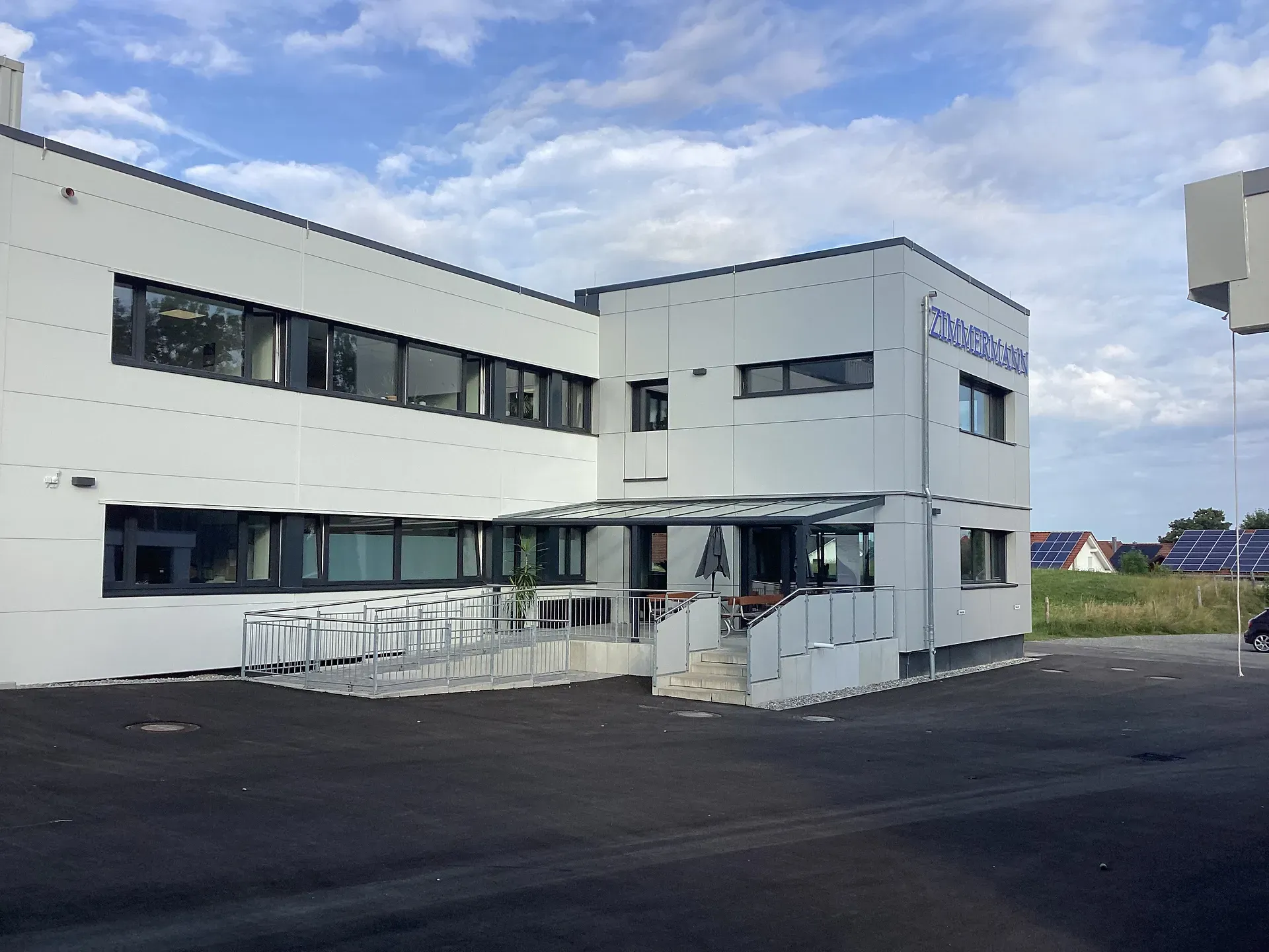 Modernes weißes Industriegebäude mit großen Fenstern, einer Rampe und einer kleinen Treppe unter einem wolkenlosen blauen Himmel.