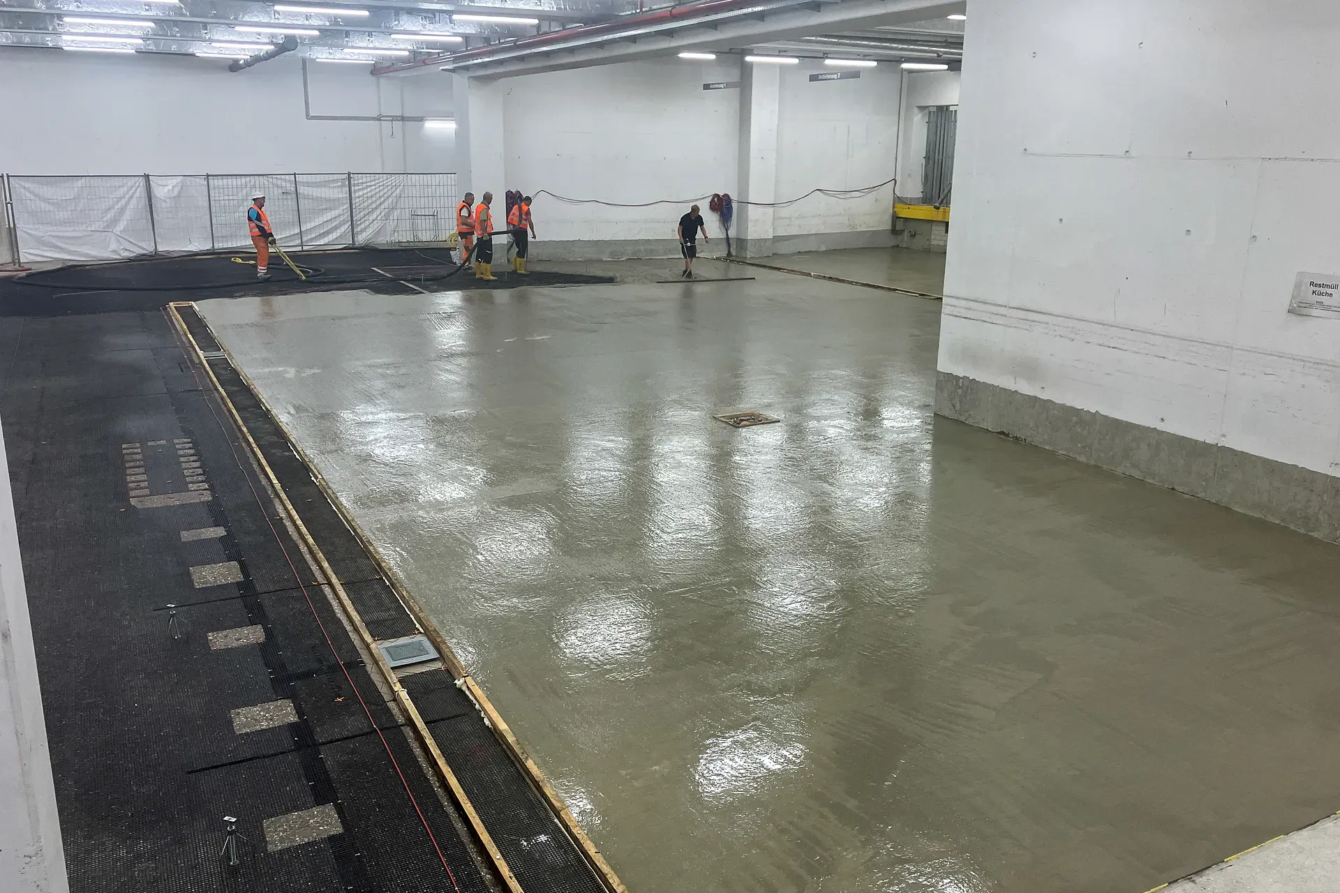 Arbeiter in Sicherheitskleidung glätten und ebnen frisch gegossenen Beton auf einem großen Boden im Innenbereich.