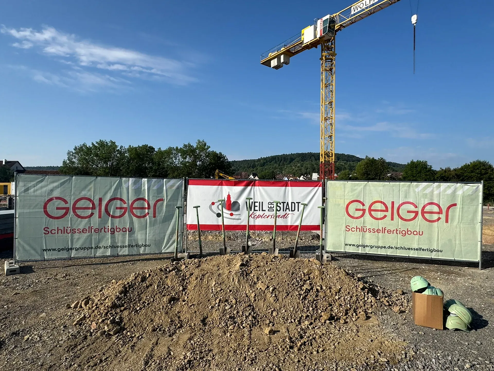 Baustelle mit den Bannern „Geiger“ und „Weil der Stadt“, einem Baukran, einem Erdhaufen und Schutzhelmen. Im Hintergrund sind Bäume zu sehen.