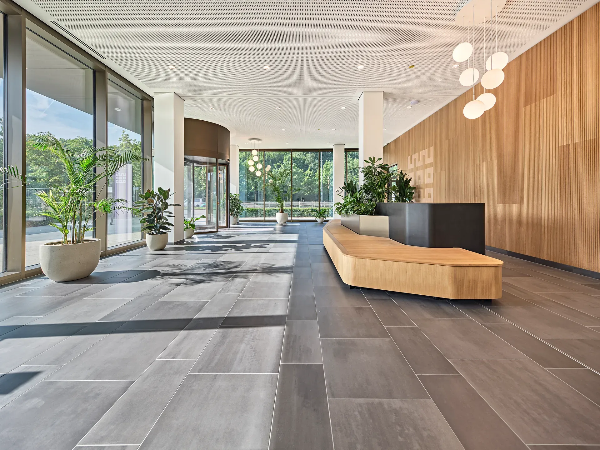 Geräumige, moderne Lobby mit großen Fenstern, grauem Fliesenboden, Akzentwand aus Holz, schwarzem Rezeptionstresen und Topfpflanzen.
