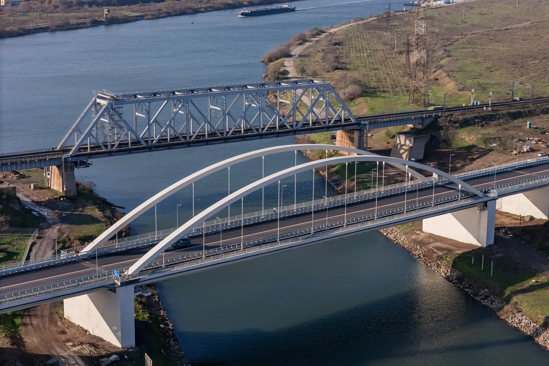 Vedere aeriană a două poduri care traversează un râu larg; unul este un pod în arc și celălalt un pod cu grinzi, înconjurate de pământ și copaci.