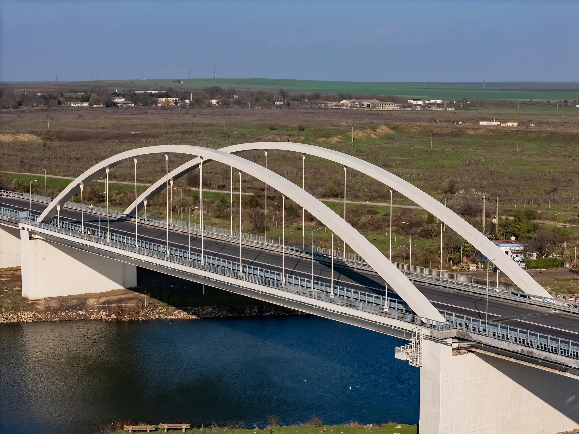 Un pod alb arcuat se întinde peste un râu liniștit, înconjurat de câmpuri verzi și clădiri împrăștiate sub un cer albastru senin.