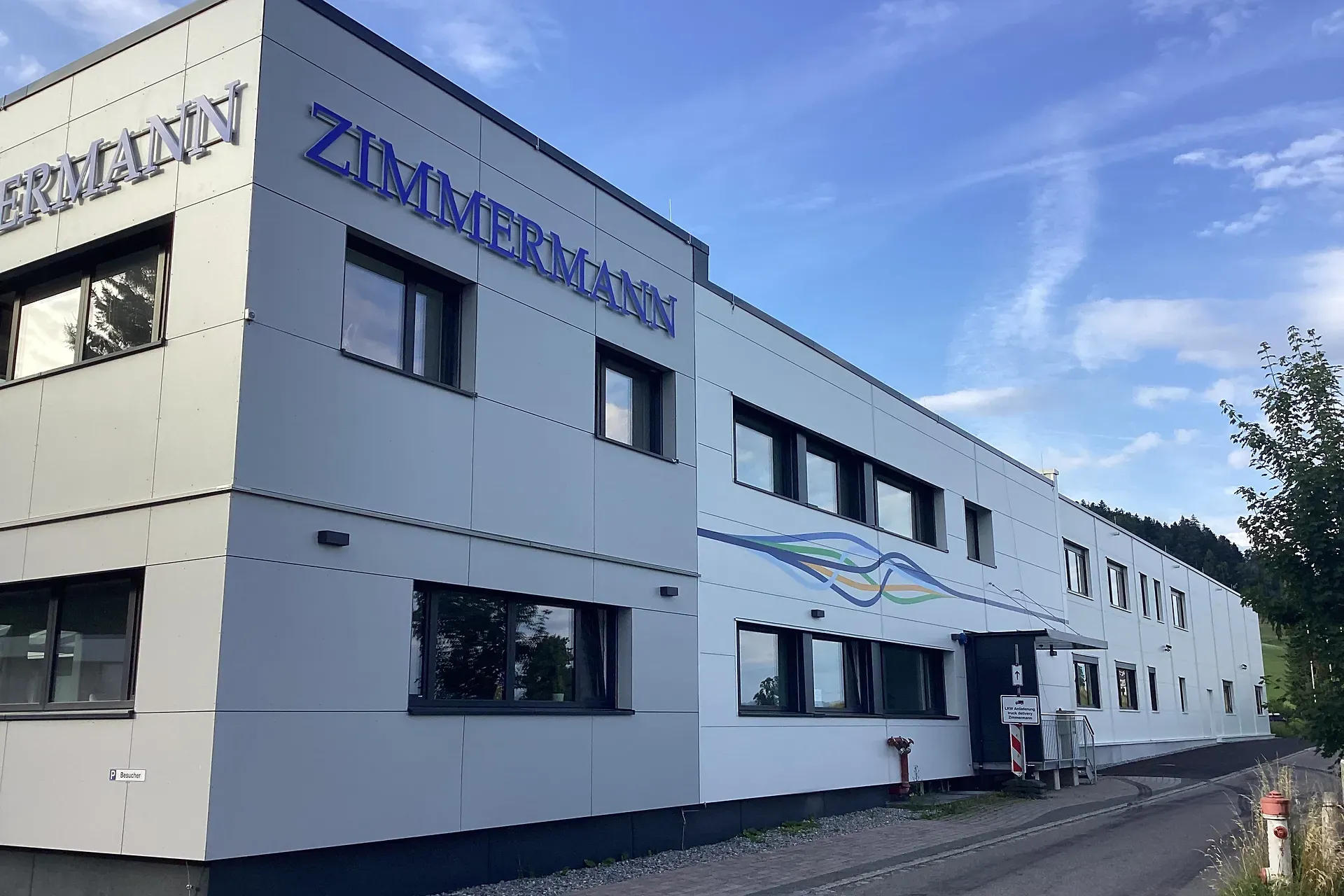 Modernes Industriegebäude mit „Zimmermann“-Beschilderung, großen Fenstern und schlichtem Design vor einem teilweise bewölkten Himmel.