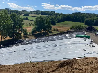 Eine Deponie mit weißer Abdeckung, im Hintergrund grüne Naturlandschaft mit schönem Wetter.