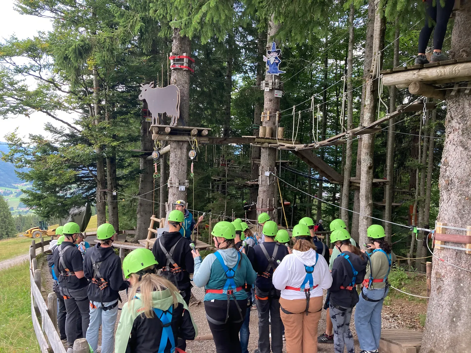 Eine Gruppe von Menschen mit grünen Helmen und Gurten versammelt sich in einem Hochseilgarten, umgeben von Bäumen und Seilrutschen.