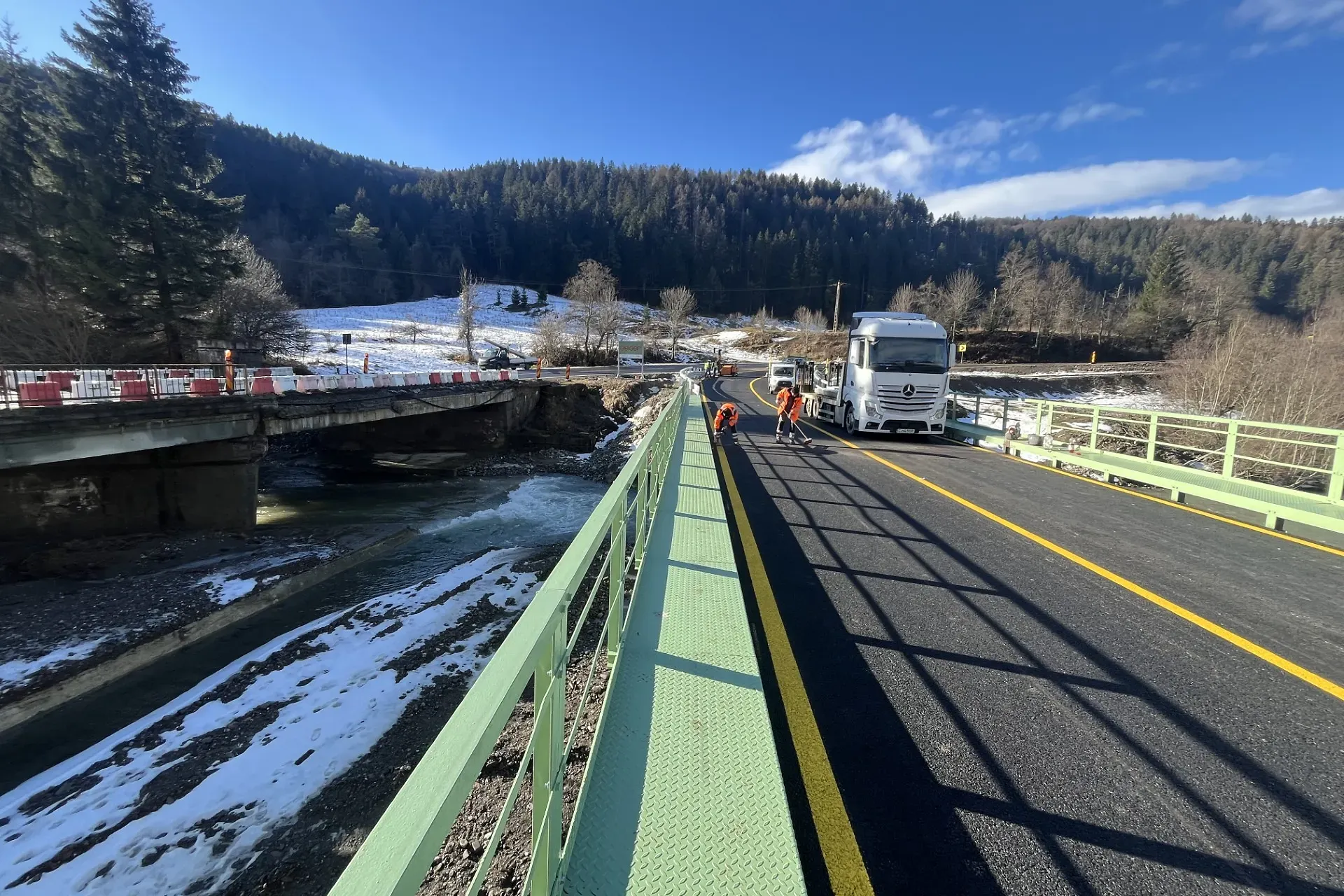 O echipă de drumuri care lucrează lângă un camion pe un pod nou, lângă unul deteriorat, înconjurați de un peisaj acoperit de zăpadă și de copaci veșnic verzi.