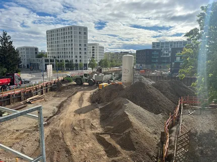 Eine Baugrube mit Bauschutt unterschiedlicher Materialien im Vordergrund. Im Hintergrund ein großes weißes Gebäude.