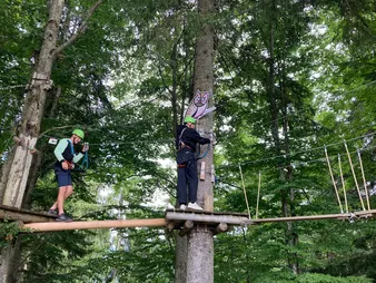 Zwei Personen mit Helmen navigieren durch einen Hochseilgarten in einem Wald. Eine Person steht auf einer Plattform und die andere auf einer Baumstammbrücke.