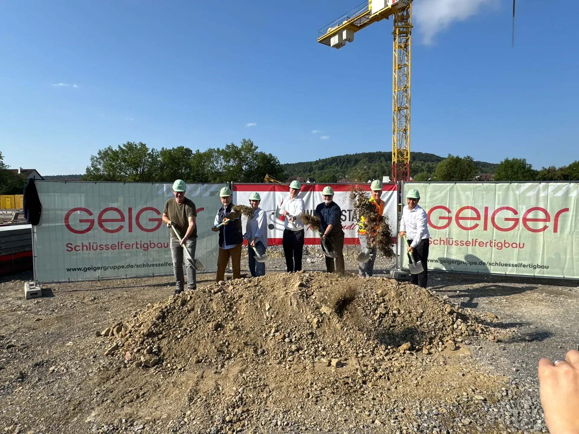 Eine Gruppe von Personen mit Schutzhelmen und Spaten nimmt an einem feierlichen Spatenstich auf einer Baustelle teil. Die Aufnahme entstand an einem sonnigen Tag.