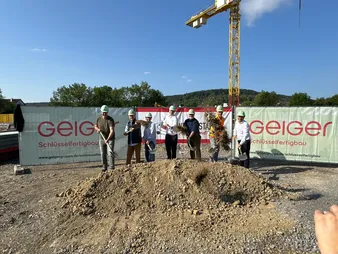 Eine Gruppe von Personen mit Schutzhelmen und Spaten nimmt an einem feierlichen Spatenstich auf einer Baustelle teil. Die Aufnahme entstand an einem sonnigen Tag.