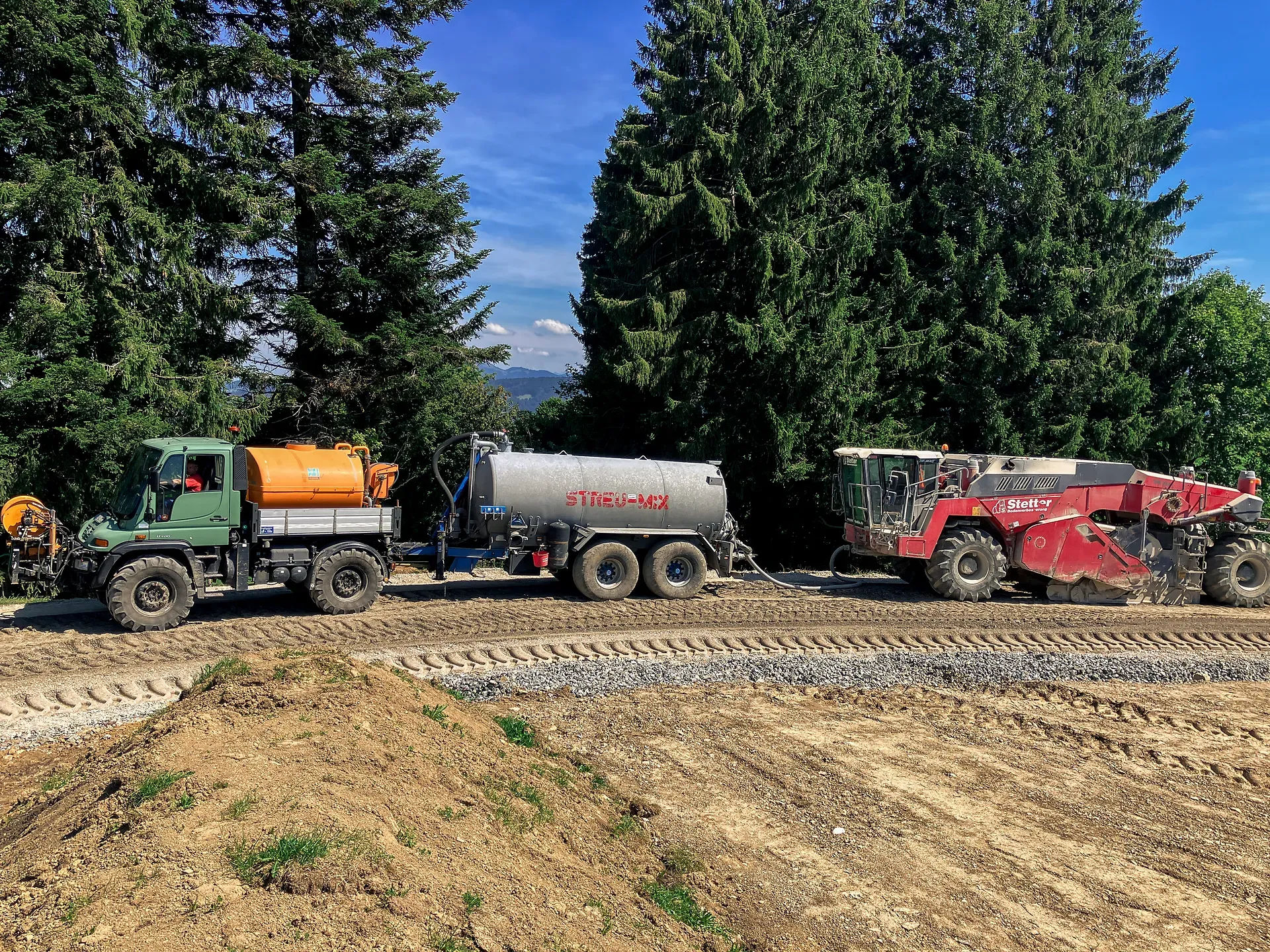 Baufahrzeuge auf einem Feldweg zwischen Bäumen; ein grüner Lastwagen, ein silberner Tankwagen und eine rote Maschine stehen aufgereiht unter einem klaren blauen Himmel.