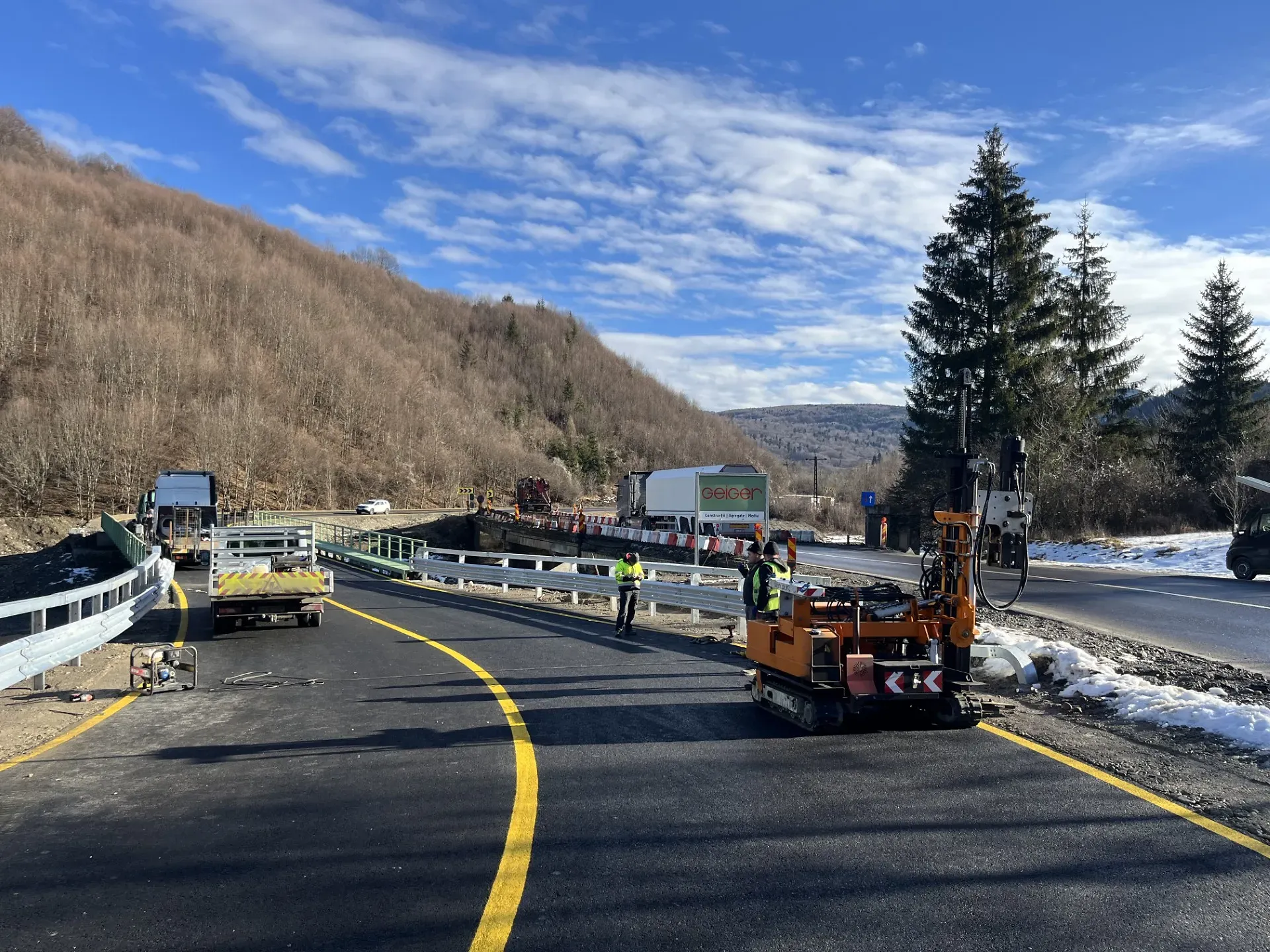 Scenă de construcție a drumului cu muncitori și utilaje pe un drum parțial asfaltat, înconjurat de copaci și dealuri sub un cer albastru senin.