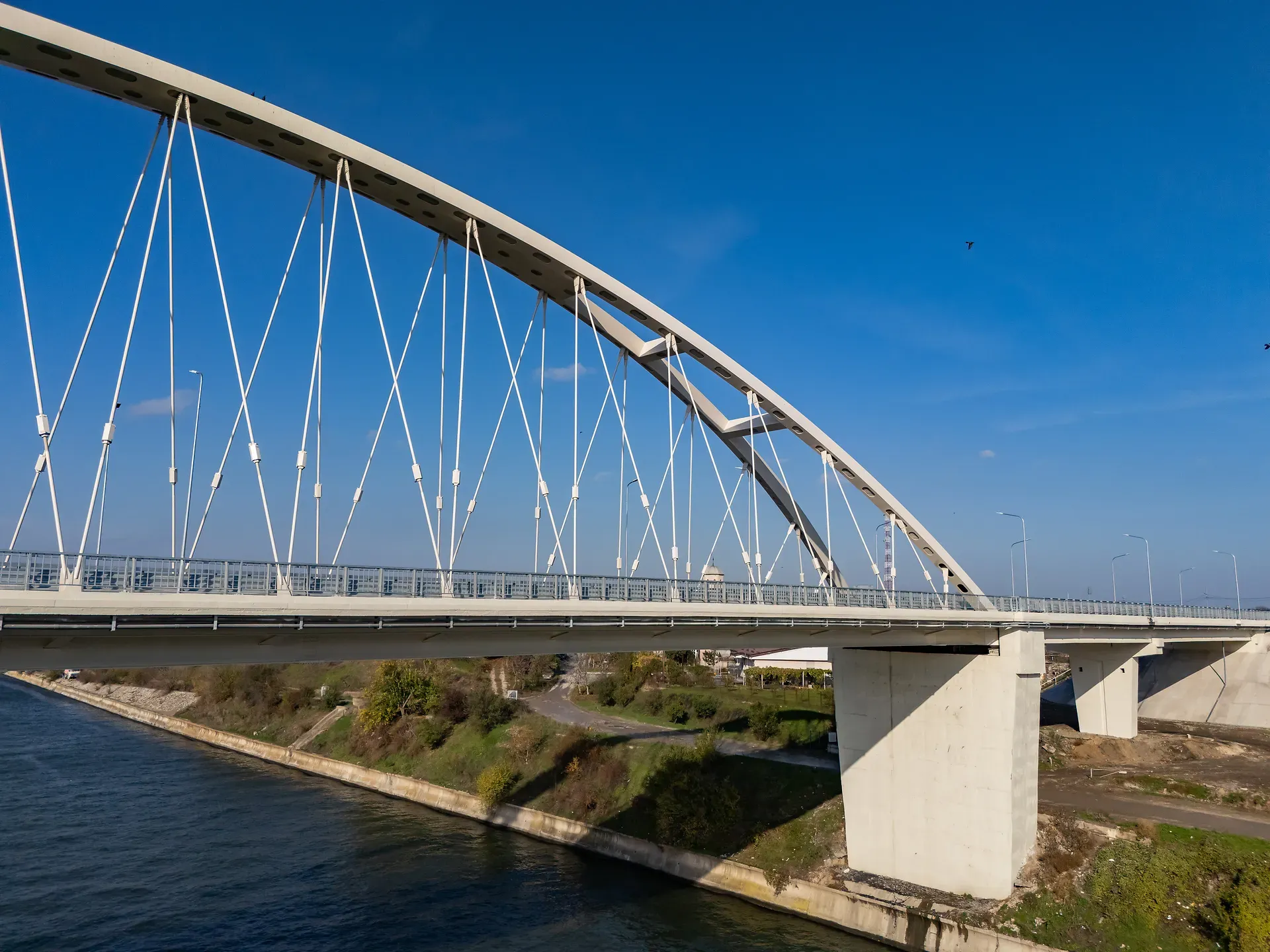 Pod arcuit cu suporturi de cablu peste un râu, cer senin albastru și peisaj verde dedesubt.