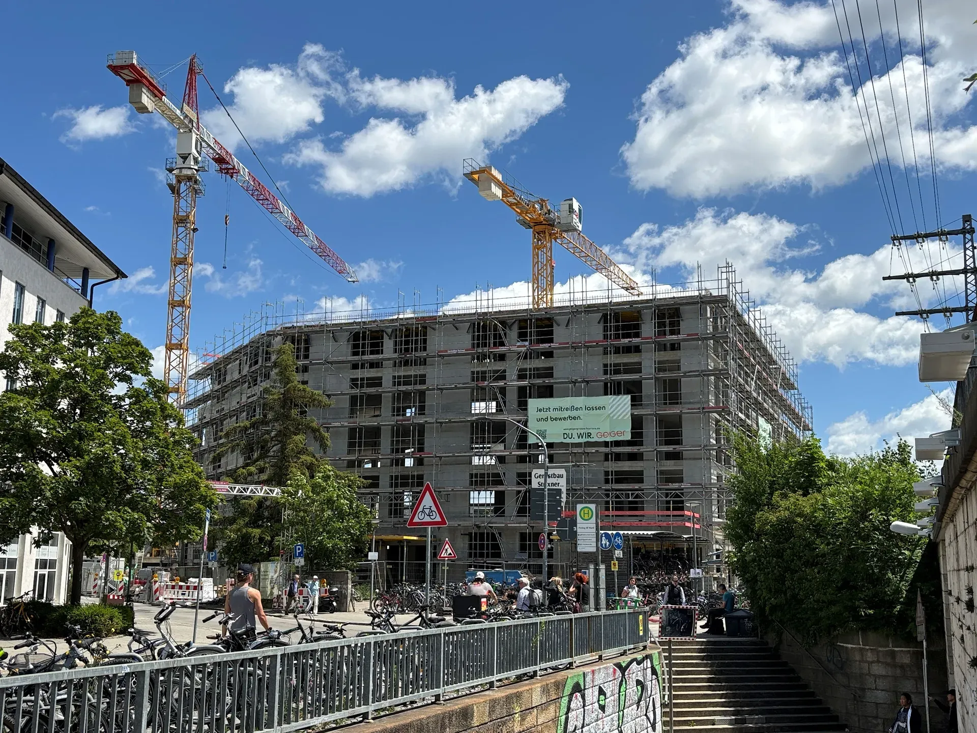 Baustelle mit Kränen und Gerüsten an einem Gebäude unter blauem Himmel. Im Vordergrund befinden sich Menschen und Fahrräder.